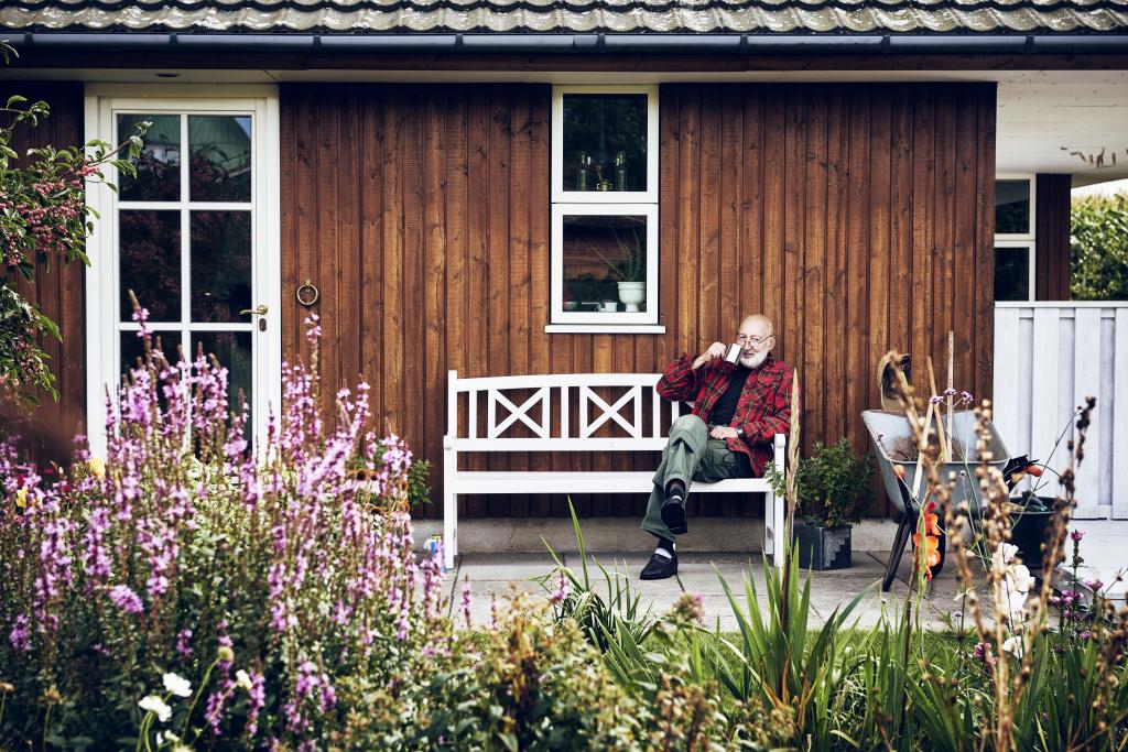 Ein Rentner sitzt auf einer weißen Bank vor einem Holzhaus und trinkt einen Kaffee.